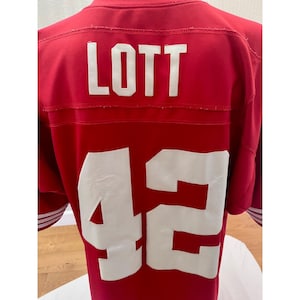 May include: A red sports jersey featuring the name LOTT in white block letters across the upper back, with the number 42 in large white digits centered below. The garment has short sleeves with white stripes and a smooth, athletic fabric finish.