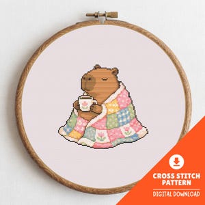 May include: Cross stitch pattern of a capybara wrapped in a patchwork quilt, holding a mug, set in a wooden embroidery hoop. The text "CROSS STITCH PATTERN DIGITAL DOWNLOAD" is in the lower right corner. A digital download.