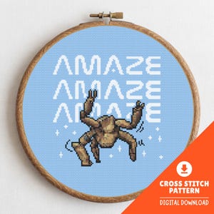May include: Cross-stitch pattern featuring a pixelated brown creature with raised arms, set against a light blue background. The word "AMAZE" is repeated three times in white. The design is framed in a wooden hoop.