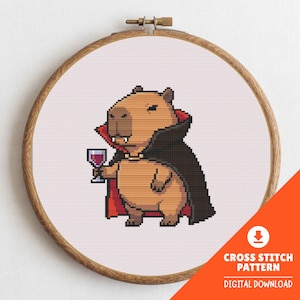 May include: A cross-stitch pattern featuring a pixelated capybara dressed as a vampire, holding a glass of red wine. The capybara wears a black cape with a red lining and a small necklace. The design is set in a wooden embroidery hoop. Text: CROSS STITCH PATTERN DIGITAL DOWNLOAD.