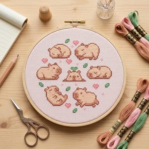 Può includere: Un telaio da ricamo a punto croce con un disegno di diversi capibara in varie pose, circondati da cuori e foglie. Il ricamo è in un telaio di legno, con filo da ricamo e altri materiali di artigianato visibili.