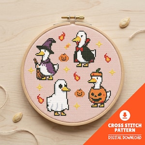 May include: A round wooden embroidery hoop containing a cross stitch pattern on light pink fabric. The design features four pixel-art ducks dressed in Halloween costumes: a witch, a vampire, a ghost, and a pumpkin, surrounded by small orange flames.