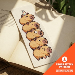 Puede incluir: Un marcapáginas rectangular con una pila vertical de cinco capibaras en pixel art bordados sobre tela Aida blanca. El capibara superior lleva un gorro de fiesta colorido. Los capibaras tienen pelaje beige, detalles marrón oscuro y expresiones faciales variadas.