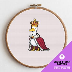 Può includere: Schema a punto croce di un'anatra pixelata con corona d'oro e mantello rosso. L'anatra è centrata su un tessuto rosa chiaro, incorniciata da un telaio da ricamo in legno. Il testo "CROSS STITCH PATTERN DIGITAL DOWNLOAD" è visibile.
