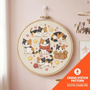 Puede incluir: Un bastidor de bordado circular de madera que contiene un patrón de punto de cruz sobre tela blanca. El diseño presenta seis gatos calicó en varias posturas, incluido uno en una caja de cartón, rodeados de corazones rosas y estrellas amarillas.