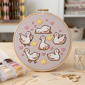 May include: A circular wooden embroidery hoop containing a cross-stitch pattern on light grey fabric. The design features six white geese in various poses, interspersed with yellow stars, pink hearts, and small letter z symbols in brown thread.