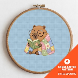May include: A cross-stitch pattern of a brown bear wearing glasses and wrapped in a patchwork quilt, reading a book. The design is set in a wooden embroidery hoop against a light blue background. The text "CROSS STITCH PATTERN DIGITAL DOWNLOAD" is in the lower right corner.