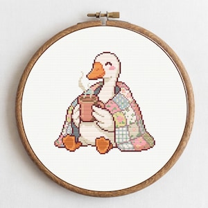 May include: Cross-stitch embroidery of a goose wrapped in a patchwork quilt, holding a steaming mug. The goose has an orange beak and feet, and is smiling. The embroidery is set in a wooden hoop.