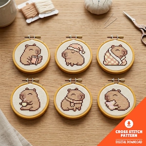 May include: Six circular wooden embroidery hoops containing cross-stitch patterns of brown capybaras in various poses, including wearing a hat, wrapped in a blanket, holding a cup, and wearing a scarf, displayed on a light wood surface.