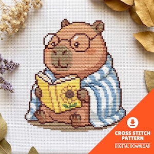 Puede incluir: Un patrón de punto de cruz que muestra un capibara marrón con gafas redondas, envuelto en una manta de rayas azules y blancas, sosteniendo un libro amarillo con una ilustración de girasol en la portada, sobre tela blanca.