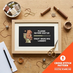 Pode incluir: Um padrão de ponto cruz emoldurado com uma capivara em uma privada, com o texto "PLEASE REMAIN SEATED GREAT IDEAS ARE LOADING". A imagem é cercada por materiais de artesanato, incluindo carretéis de linha, tesouras e agulhas. Download digital disponível.