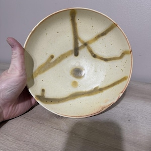 May include: A handmade ceramic bowl with a cream-colored interior and a reddish-brown rim. The bowl features brown, abstract linear designs. The bowl is held by a person.