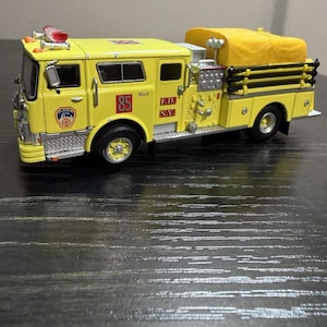 May include: A yellow toy fire engine with the number 85 on the side. The words "Mack" and "F.D. N.Y." are printed on the side. The engine has a yellow tarp covering the back and red lights on top.
