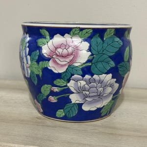 May include: A blue ceramic planter with a floral design. The planter features large pink and white flowers with green leaves against a dark blue background. The planter is a round shape.