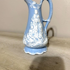 May include: A light blue ceramic pitcher with a white relief design of figures. The pitcher has a curved handle and a spout. The base is a scalloped design. The pitcher is approximately 6 inches tall.