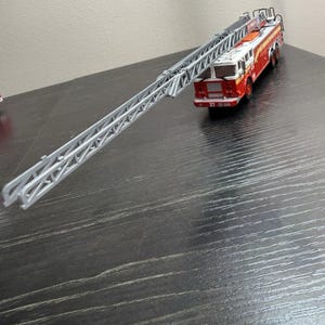 May include: A red and white toy fire engine with an extended silver ladder. The vehicle has black wheels and is on a dark surface. The ladder extends diagonally across the frame.