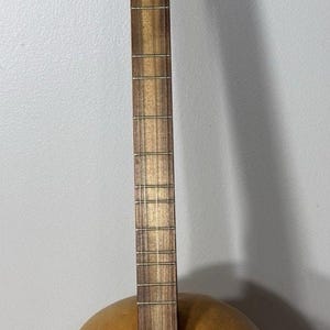 May include: A stringed instrument with a round, gourd-shaped body and a long neck. The body is a light brown color, and the neck is made of wood. The instrument has strings and tuning pegs.