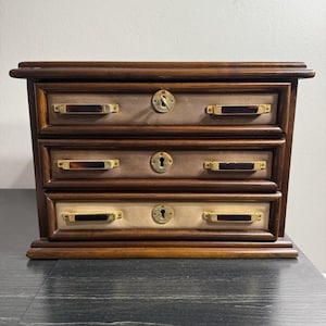 Op de afbeelding: Een kleine houten sieradendoos met drie lades en een donkerbruine afwerking. Elke lade heeft een messingkleurige handgreep en sleutelgat. De ladefronten zijn lichtbruin met een beige middenstuk. De doos staat op een zwart oppervlak.