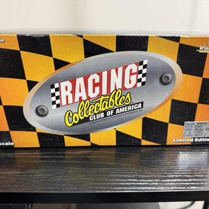 Op de afbeelding: Een Racing Collectables Club of America doos met een zwart-geel geblokt patroon. De doos heeft de tekst "RACING Collectables" en "Winston Cup". Het is een verzamelobject op schaal 1:24 in beperkte oplage.