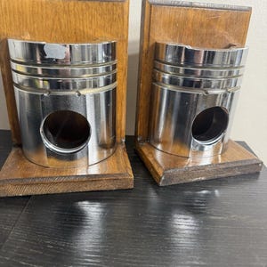 Könnte beinhalten: Zwei einzigartige Buchstützen mit polierten Metallkolben, die auf gebeizten Holzsockeln montiert sind. Die Kolben haben eine zylindrische Form mit einer großen kreisförmigen Öffnung. Das Holz hat eine natürliche Maserung und einen warmen, braunen Farbton.