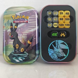 Può includere: Una scatola di metallo del gioco di carte collezionabili Pokemon con un'illustrazione di Umbreon e una scatola aperta abbinata contenente dadi danno gialli e neri numerati, due dadi neri speciali e una moneta blu circolare.
