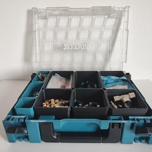 Peut inclure: Une mallette de rangement en plastique bleu sarcelle et noir avec un couvercle transparent à charnières portant le logo Makita. L'intérieur contient plusieurs bacs amovibles remplis de divers composants de quincaillerie.