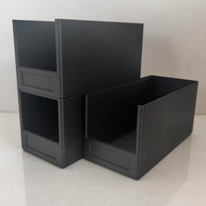 Könnte beinhalten: Drei mattschwarze Kunststoff-Aufbewahrungsboxen mit offener Vorderseite. Zwei Boxen sind auf der linken Seite vertikal gestapelt, während eine Box separat rechts steht. Jede Box verfügt über ein rechteckiges, vertieftes Etikettenfeld vorne.