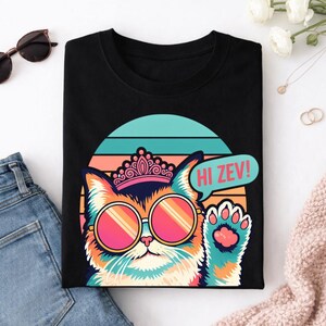 Könnte beinhalten: Schwarzes T-Shirt mit einer Comic-Katze, die eine Sonnenbrille und eine Krone trägt. Die Katze winkt mit einer Sprechblase mit der Aufschrift "HI ZEV!". Das Design hat einen Retro-Sonnenuntergangshintergrund.