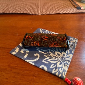 Könnte beinhalten: Eine rechteckige, schwarz-braune dekorative Box mit roten Akzenten liegt auf einem blau gemusterten Stoff. Die Box hat eine glänzende Oberfläche. Eine rot-weiße Quaste hängt an der Ecke des Stoffes.