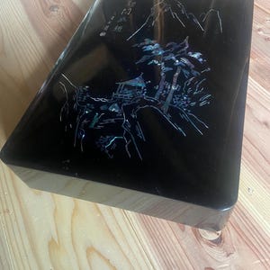 以下が含まれることがあります： 長方形の黒い漆塗りの箱。蓋には、山、木、建物が描かれた詳細な風景画が、虹色の象嵌で描かれています。箱は木製の表面に置かれています。