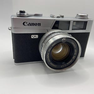 May include: A vintage Canon QL camera with a silver top and black body. The lens is silver with the text "Canon Lens SE 45mm 1:1.7". The word "Canon" is printed on the top of the camera.