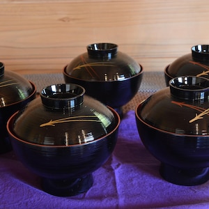 May include: Five black lacquered Japanese soup bowls with lids. Each bowl has a red rim and a gold and brown arrow design on the lid. The bowls are on a purple cloth.