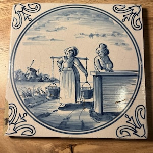 May include: A square ceramic tile featuring a blue and white hand-painted scene inside a circular border. The illustration depicts a person carrying two buckets on a yoke, a second person leaning on a structure, and a windmill in the background.