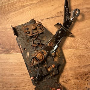 Op de afbeelding: Een vintage Hurst auto-versnellingspook met een zwarte hendel waarop het woord HURST in het wit is gedrukt. Het mechanisme bevat een verroeste metalen basisplaat met diverse veren, schroeven en elektrische bedradingscomponenten bevestigd.