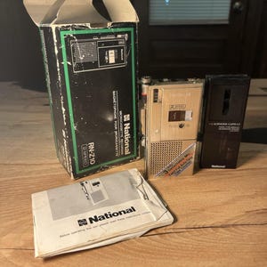 May include: A National RN-Z10 microcassette recorder in a gold finish, accompanied by its original box, a black microphone capsule, and an instruction manual. The recorder features a 2-speed setting, a tape counter, and a detachable microphone.