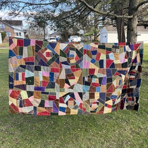 Può includere: Una grande trapunta rettangolare caratterizzata da un patchwork caotico di ritagli di tessuto multicolore in tonalità di rosso, blu, verde, giallo e marrone. I pezzi sono uniti da spesse cuciture a zigzag chiare visibili sulla superficie.