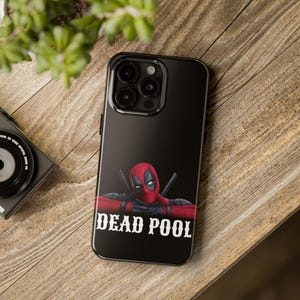 May include: Black phone case with a Deadpool design. The case features the character in a red and black costume, with the text "DEAD POOL" in white. The phone case is on a wooden surface with a camera and plant.