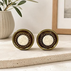 Op de afbeelding: Twee ronde kastknoppen met een centraal gebroken wit gespikkeld inlegstuk, omringd door een textuurrijk antiek messingkleurig metalen frame met een kralenrand. De knoppen hebben een glad, afgerond profiel en een metalen afwerking.