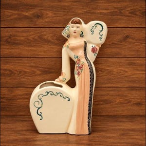 May include: A cream-colored ceramic wall pocket vase shaped like a standing figure wearing a peach-colored gown with red floral accents and a black decorative trim. The figure holds a large, curved, cream-colored vessel with green scrollwork.