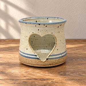 May include: A speckled ceramic candle holder with a heart-shaped cutout on the front. The piece features a cream-colored base with dark brown speckles, accented by two horizontal blue stripes near the bottom and a blue rim around the top opening.