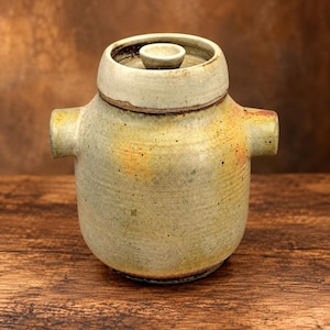 Op de afbeelding: Een cilindrische keramische pot met een bijpassend deksel voorzien van een kleine centrale knop. De pot heeft twee uitstekende zijhandvatten en een gespikkeld glazuur in tinten beige, crème en gedempt geel met subtiele donkere spikkels.