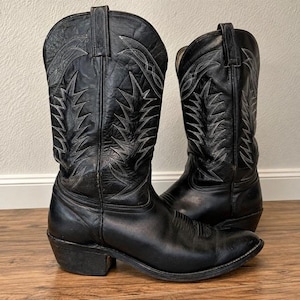 Puede incluir: Un par de botas vaqueras de cuero negro con una caña alta que presenta intrincadas costuras grises en un patrón dentado similar a llamas. Las botas tienen punta afilada, tacón apilado y tiradores en la parte superior de la caña, diseño clásico.