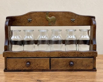 Puede incluir: Un especiero de madera marrón oscuro con dos cajones pequeños con pomos redondos, seis botellas de vidrio transparente y un panel trasero decorativo adornado con un emblema central de gallo de latón y dos estrellas.