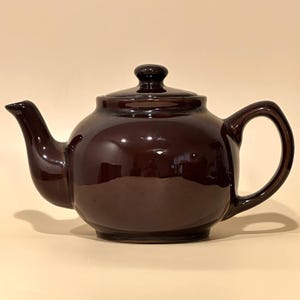 May include: A glossy, dark brown ceramic teapot with a rounded body, a curved spout, a sturdy handle, and a matching lid with a small knob handle on top. The surface of the teapot is smooth and reflective, showing light highlights across its exterior.