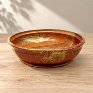 Könnte beinhalten: Eine flache, runde Keramikschale mit einer variierten Glasur in tiefen Orange-, Rost- und Erdbrauntönen. Die Oberfläche weist eine gesprenkelte Textur mit subtilen gelben und grünen Akzenten entlang des Randes und des inneren Bodens auf.