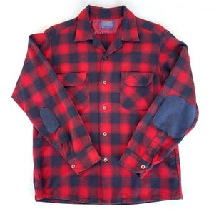 May include: Red and black checked flannel shirt with navy blue elbow patches. The shirt features two chest pockets and a button-down front. The collar is a classic spread collar style.