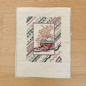 Peut inclure: Un morceau rectangulaire de tissu en lin blanc cassé présentant une broderie au point de croix centrale d'un vase à motifs rempli de fleurs roses. Le motif est encadré par une bordure géométrique dans des tons de vert, rose, bleu et gris.