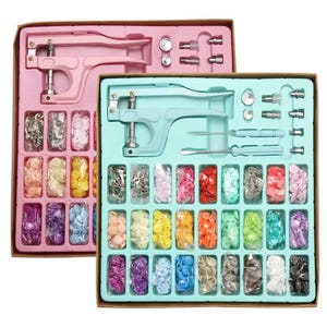 May include: Two rectangular cardboard boxes containing plastic snap fastener kits. Each kit includes a metal plier tool, various metal dies, a screwdriver, and numerous small plastic bags filled with colorful snap buttons in a variety of shades.