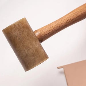 Puede incluir: Un mazo con una cabeza cilíndrica, translúcida y de color marrón claro con una superficie texturizada y un mango de madera lisa de color claro. La herramienta está colocada en diagonal sobre un fondo liso y claro, mostrando la cabeza y el mango.
