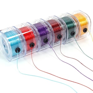 Könnte beinhalten: Ein Set aus sechs Spulen mit farbiger Nylonschnur in einem transparenten Kunststoff-Spendergehäuse. Die Spulen haben leuchtende Farben in Hellblau, Rot, Violett, Dunkelrot, Dunkelgrün und Gelb, jeweils mit einer schwarzen Öse zur Führung.